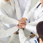 View looking down on five physicians with hands reaching into center of a circle symbolizing consensus